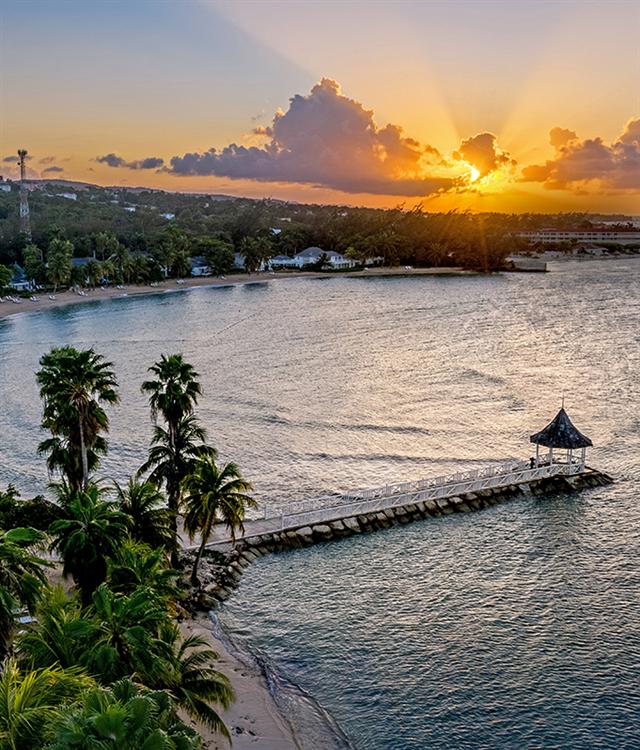 A dock over water with palm trees and a beach and a sunset